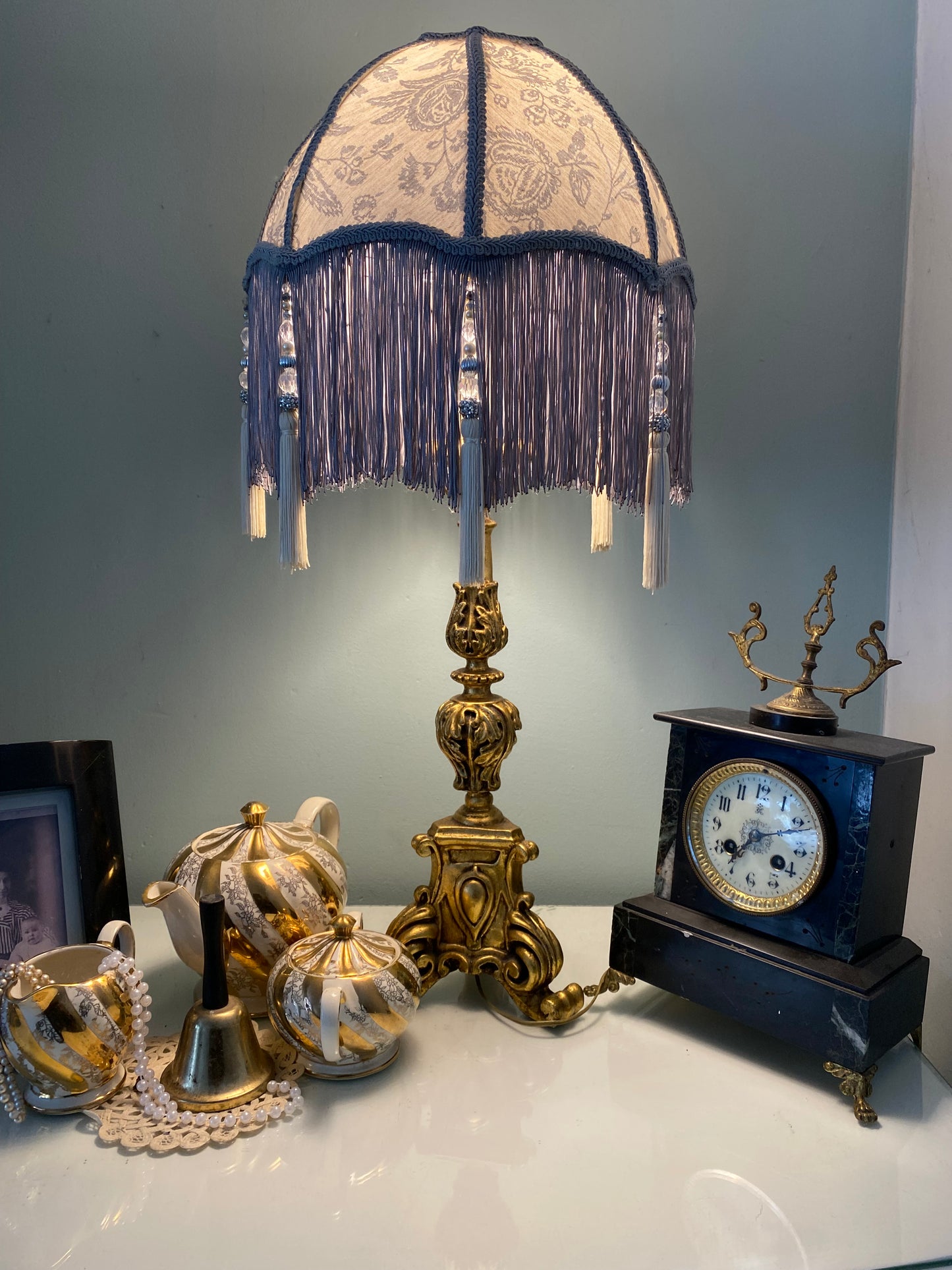 Full lampshade view – A grey Victorian parachute-style lampshade in blue toile fabric, with a soft scalloped shape and a long beaded fringe.Lit up ambient light