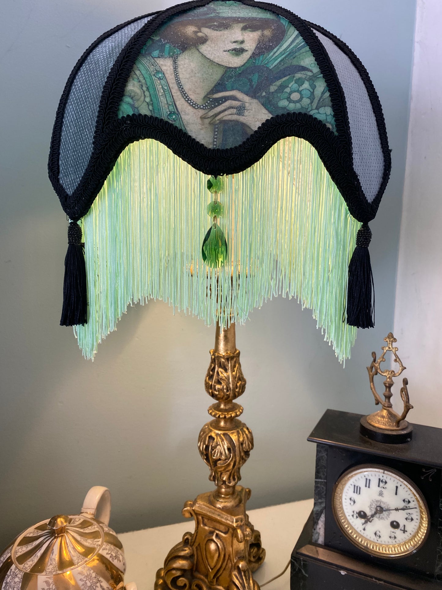 Lit lamp / ambience shot – “Victorian parlour lampshade illuminated to reveal Edwardian lady image, emerald jewels, and glowing green fringe – antique-inspired decorative lighting.”front view