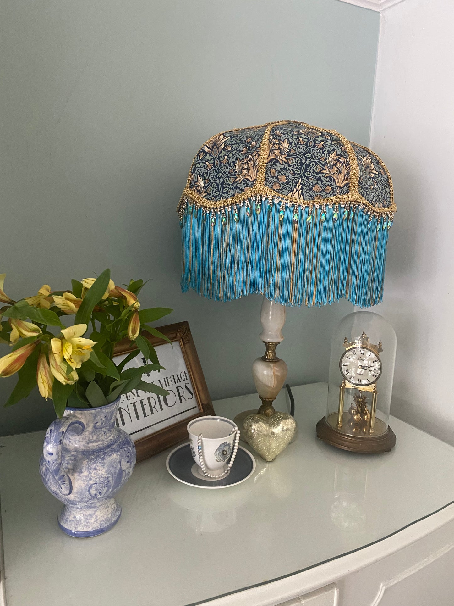 Angled shot, unlit
Handmade Art Deco lampshade in William Morris “Snakeshead” turquoise fabric, shown unlit at an angle to highlight the gold and turquoise two-tone fringe, gold braid trim, and iridescent diamanté detail — elegant Victorian-inspired fringed lighting.vintage setting
