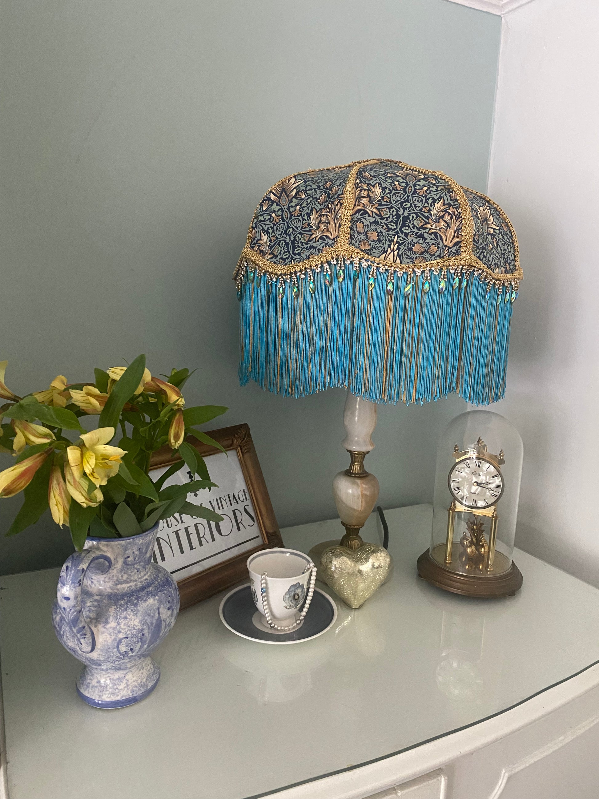 Angled shot, unlit
Handmade Art Deco lampshade in William Morris “Snakeshead” turquoise fabric, shown unlit at an angle to highlight the gold and turquoise two-tone fringe, gold braid trim, and iridescent diamanté detail — elegant Victorian-inspired fringed lighting.vintage setting