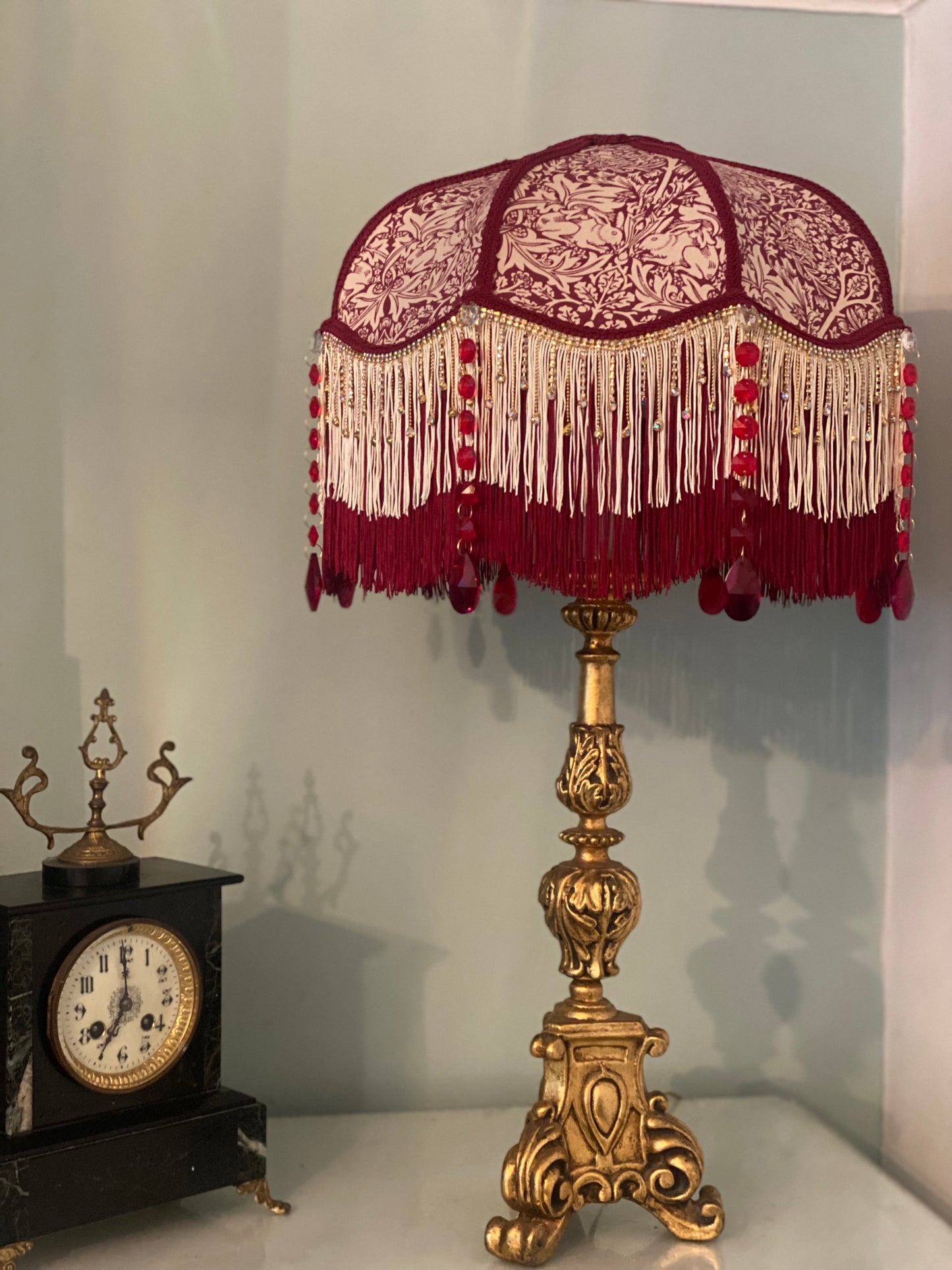 Full view – “Handmade Victorian lampshade in William Morris ‘Brer Rabbit’ fabric with burgundy trim, gold diamante fringe, and ruby glass crystal gems – elegant Arts and Crafts inspired lighting.” with antique clock