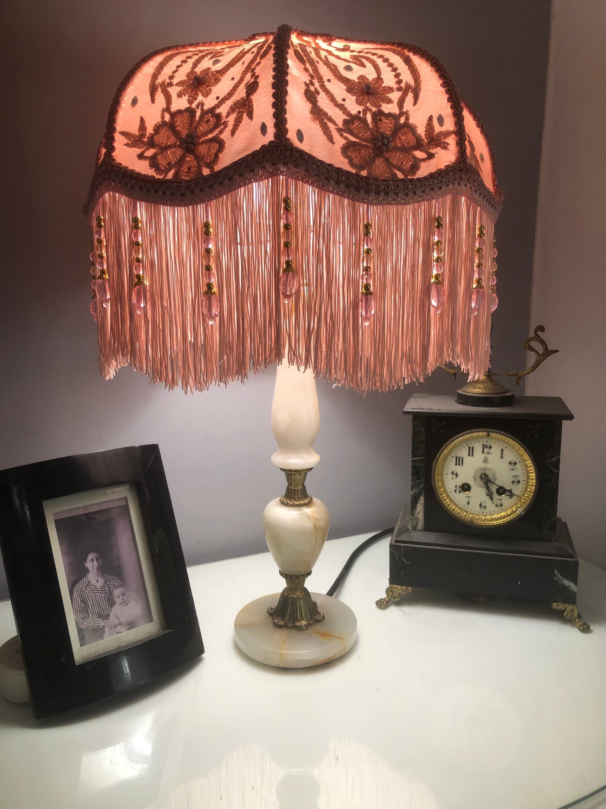 Lit lifestyle setting – “Handmade pink Victorian silk lampshade with floral overlay and pink fringe, glowing softly in a vintage-inspired interior.”