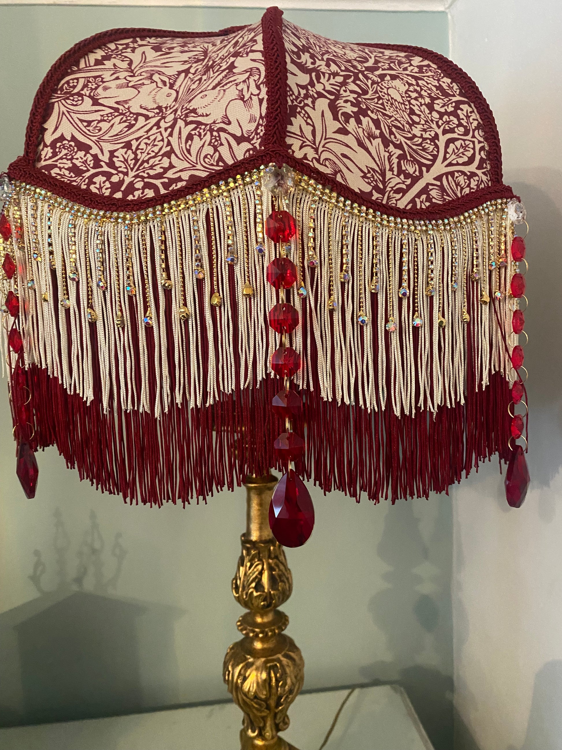 ambience shot – “William Morris ‘Brer Rabbit’ Victorian lampshade illuminated, revealing gold fringe, ruby crystals, and warm Arts and Crafts style glow.”