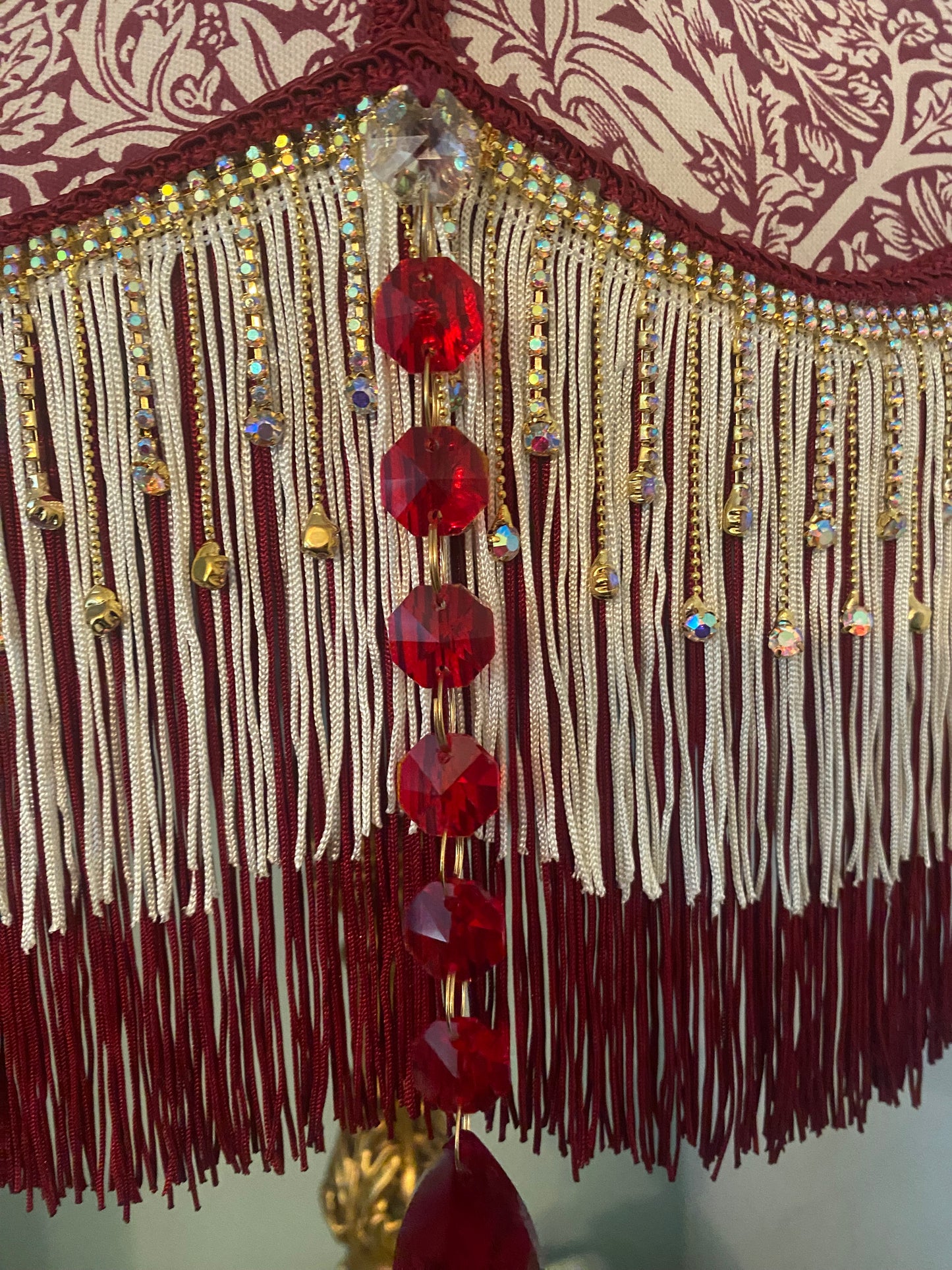 Close-up of ruby crystal gems – “Ruby red glass crystal gems sparkling along the gold fringe of a William Morris ‘Brer Rabbit’ Victorian lampshade – luxurious handmade lighting accent.”